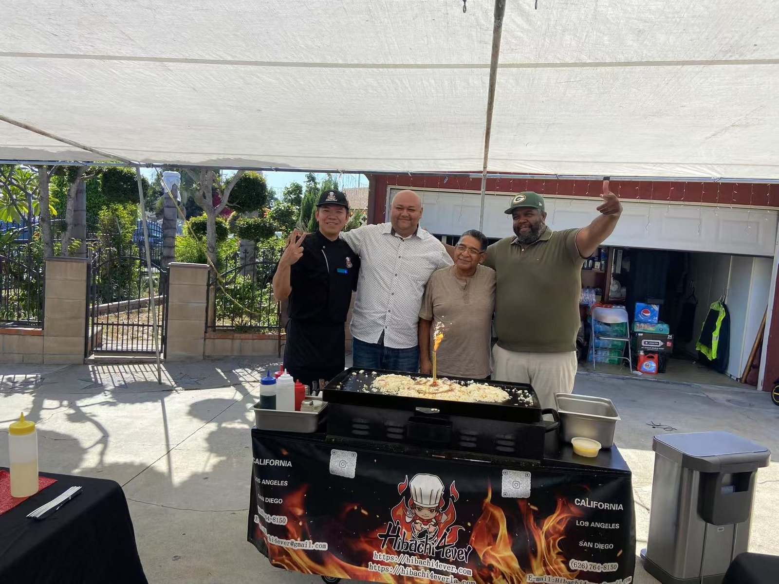 Four people stand smiling behind a hibachi grill under a canopy in a Los Angeles driveway, with cooking supplies and a garage in the background.