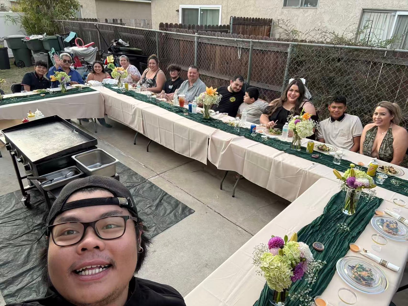 A group of people sit around two long tables set for a meal in a Los Angeles backyard, enjoying what looks like a lively hibachi gathering. In the foreground, someone snaps a selfie with the group.